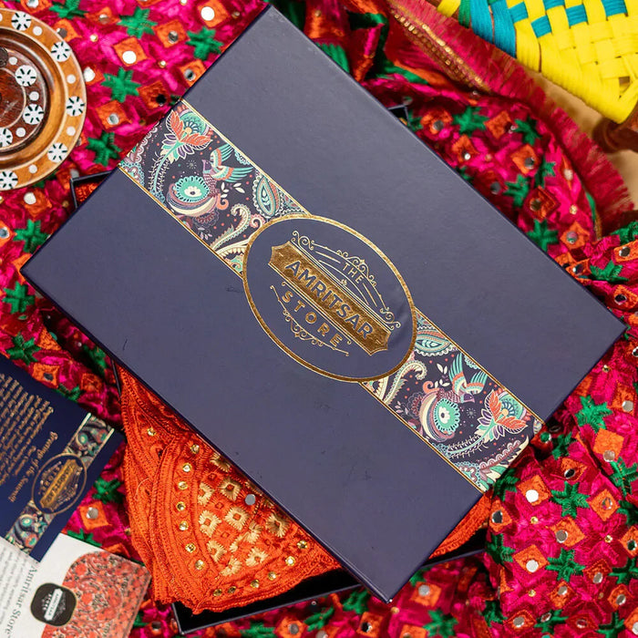 A dark navy blue The Amritsar Store gift box with ornate gold and multicolor patterned detailing, presented on a bed of vibrant, colorful hand embroidered textiles.