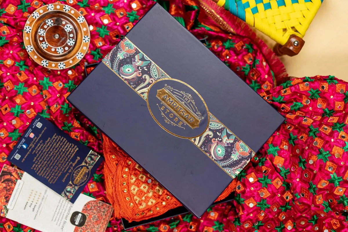 A luxury gift box set from The Amritsar Store, featuring a navy blue main box with a gold and multicolor paisley band, surrounded by traditional Indian textiles.